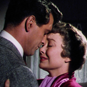 A man in a suit and a woman in a pink dress stand close, sharing an intimate moment against a softly lit backdrop.