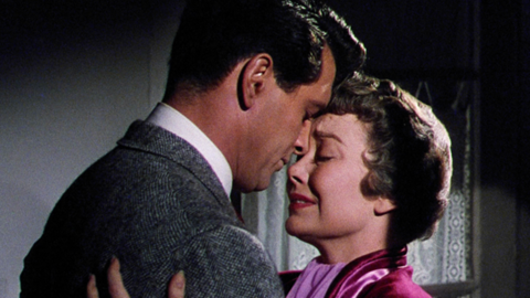 A man in a suit and a woman in a pink dress stand close, sharing an intimate moment against a softly lit backdrop.
