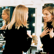 A person with long blonde hair wearing a black top is reflected in a vanity mirror, surrounded by lights and personal photos.