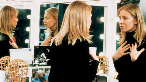 A person with long blonde hair wearing a black top is reflected in a vanity mirror, surrounded by lights and personal photos.