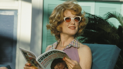 A still from the film Far From Heaven. A woman in a purple dress sits on a lounge chair, reading a Cosmopolitan magazine.