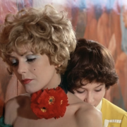 Two women embrace each other against a vibrant floral backdrop, showcasing distinct hairstyles and colorful attire.