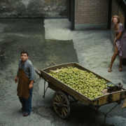 A man in a checkered shirt and apron stands beside a wooden cart filled with green pears in a narrow alley. A woman peeks from a doorway.