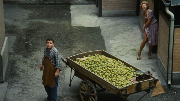 A man in a checkered shirt and apron stands beside a wooden cart filled with green pears in a narrow alley. A woman peeks from a doorway.