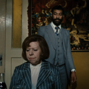 A woman in a striped suit sits with a drink in front of a vintage tapestry, while a man in a gray suit stands behind her.