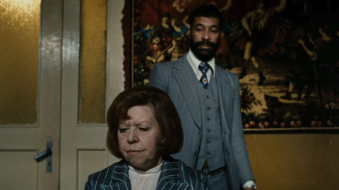 A woman in a striped suit sits with a drink in front of a vintage tapestry, while a man in a gray suit stands behind her.