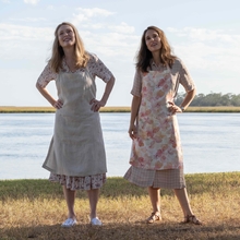Two women stand outdoors by a river, dressed in aprons, with a tree trunk beside them and a scenic landscape in the background.
