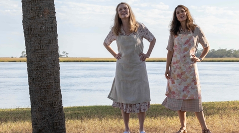 Two women stand outdoors by a river, dressed in aprons, with a tree trunk beside them and a scenic landscape in the background.