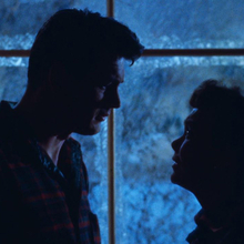 Silhouettes of a couple standing close together, framed by frosted windows, conveying a moment of intimacy in a cold, dimly lit setting.