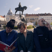 A still from the film Kontinental '25 - five people sit on a bench, two of them looking at a book, while the others are drinking and smoking.