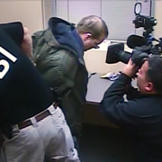 A KBI agent detains a person in a small room while a cameraman records the scene.