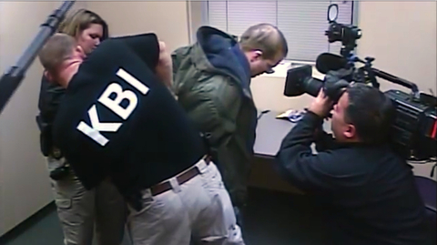 A KBI agent detains a person in a small room while a cameraman records the scene.