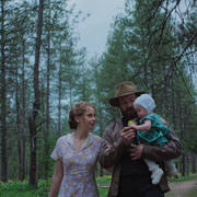 A woman and a man stroll through a pine forest, holding a child in a green dress, surrounded by lush greenery.