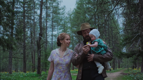A woman and a man stroll through a pine forest, holding a child in a green dress, surrounded by lush greenery.