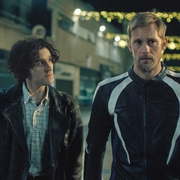 Two men stand side by side on a dimly lit street adorned with decorative lights.