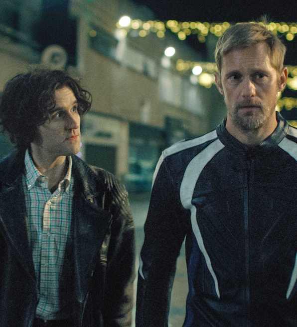 Two men stand side by side on a dimly lit street adorned with decorative lights.