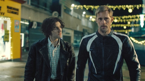 Two men stand side by side on a dimly lit street adorned with decorative lights.