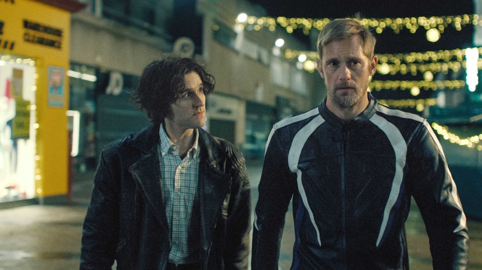 Two men stand side by side on a dimly lit street adorned with decorative lights.