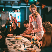 A person serves a colorful layered cake at a festive gathering, surrounded by guests engaged in conversation.