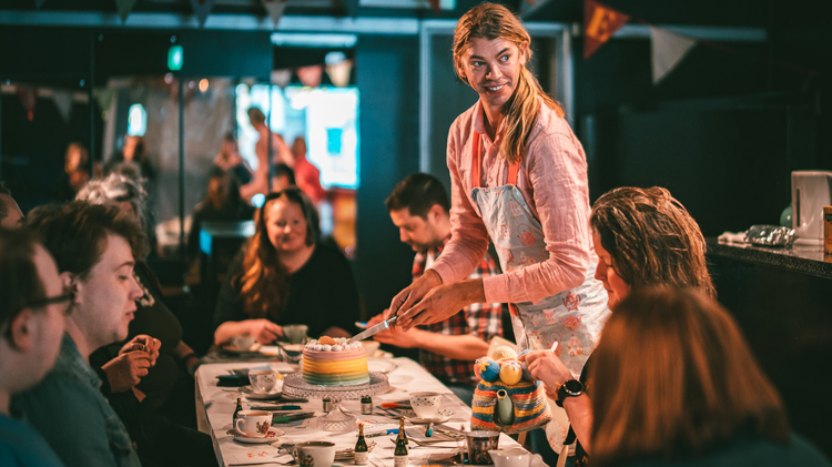 A person serves a colorful layered cake at a festive gathering, surrounded by guests engaged in conversation.