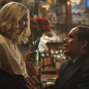 A woman in a light blouse holds a bouquet of red flowers, while a man in a dark suit gestures expressively in a bar setting.