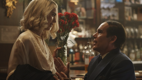 A woman in a light blouse holds a bouquet of red flowers, while a man in a dark suit gestures expressively in a bar setting.