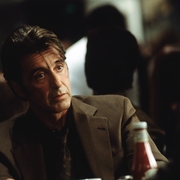 A man in a brown suit sits at a table, with a bottle of ketchup visible, amidst a blurred crowd in a restaurant setting.