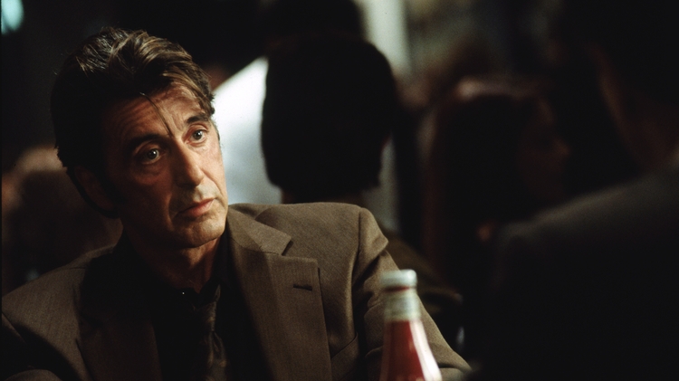 A man in a brown suit sits at a table, with a bottle of ketchup visible, amidst a blurred crowd in a restaurant setting.