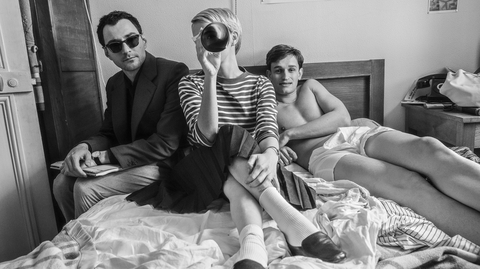 A woman in a striped shirt and skirt holds a bottle, seated on a bed with two men, amidst scattered blankets and casual decor.