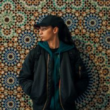 A person in a black jacket and green hoodie stands against a vibrant, intricate patterned tile wall.