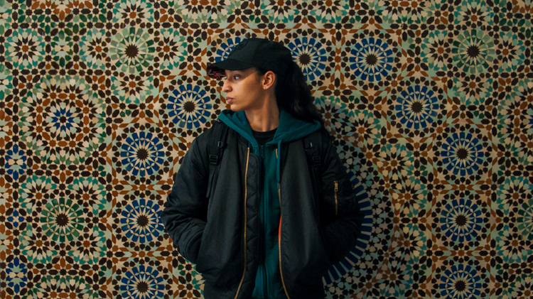 A person in a black jacket and green hoodie stands against a vibrant, intricate patterned tile wall.