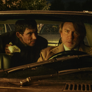 Two individuals are engaged in a tense conversation inside a parked car, with raindrops on the windshield and a dark, moody atmosphere.