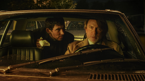Two individuals are engaged in a tense conversation inside a parked car, with raindrops on the windshield and a dark, moody atmosphere.