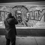 A person photographs a vibrant mural of George Floyd, featuring names of other victims of racial violence, expressing solidarity and remembrance.