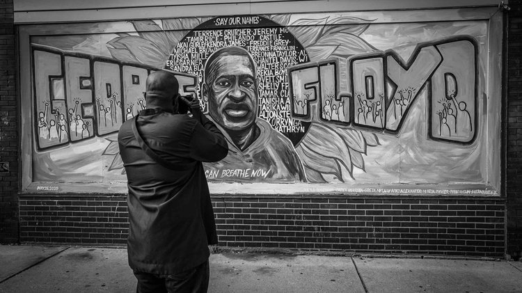 A person photographs a vibrant mural of George Floyd, featuring names of other victims of racial violence, expressing solidarity and remembrance.