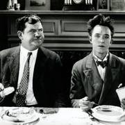 Two men dressed in formal attire sit at a dining table with plates of food in a vintage setting.