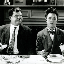 Two men dressed in formal attire sit at a dining table with plates of food in a vintage setting.