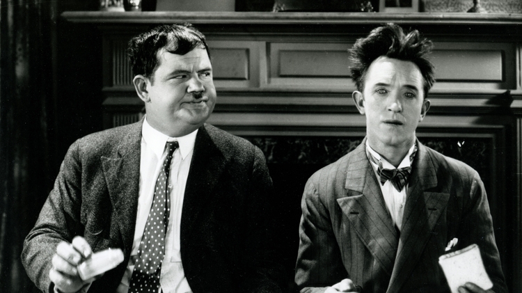 Two men dressed in formal attire sit at a dining table with plates of food in a vintage setting.