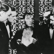 An gathering of elegantly dressed individuals, against an ornate ironwork background.