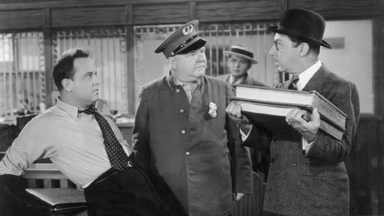 A man in a polka dot tie leans on a desk while two other men, one in a police uniform and another in a suit, engage in conversation.