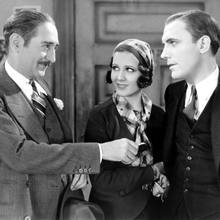 Three elegantly dressed individuals engage in conversation, with one holding a small object in their hand against a vintage backdrop.