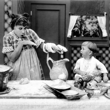 A woman in an apron playfully holds a dough snake while a child sits beside her, surrounded by various cooking utensils and ingredients.
