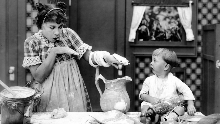 A woman in an apron playfully holds a dough snake while a child sits beside her, surrounded by various cooking utensils and ingredients.