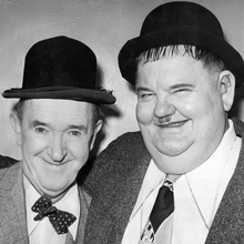 Two men in vintage attire, wearing bowler hats, posing closely together against a simple background. One has a polka dot bow tie.