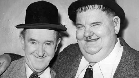 Two men in vintage attire, wearing bowler hats, posing closely together against a simple background. One has a polka dot bow tie.