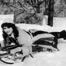 A woman lying down on a sledge is smiling in a snowy scene.