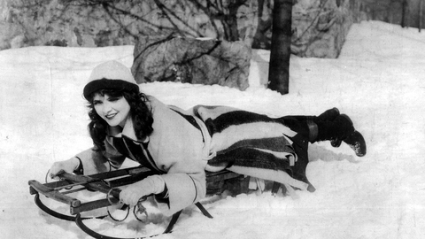 A woman lying down on a sledge is smiling in a snowy scene.