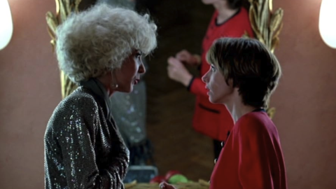 Two women engage in conversation in front of a vintage mirror, accentuated by elegant lighting and retro-style decor.