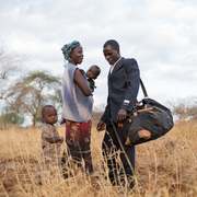 A man in a suit stands with a woman holding a child, while a young child looks on in a dry, grassy field.