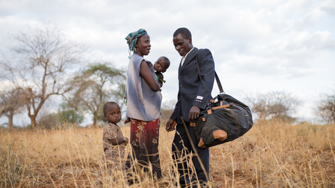 A man in a suit stands with a woman holding a child, while a young child looks on in a dry, grassy field.